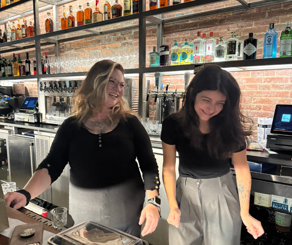 two women smiling and laughing behind a cocktail bar